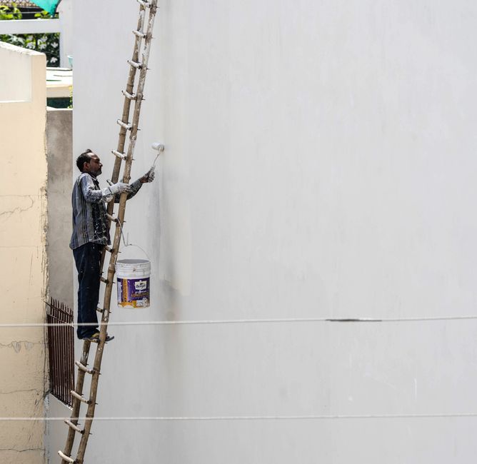 A painter on a ladder applying paint to a building's exterior wall with a roller brush outdoors in summer heat.